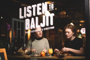 To jenter/damer spiser i vinduet i restauranten. De snakker med hverandre. Vi ser et rødvinn glass og et glass med mango lassi. Listen to Baljit logo i vindu med hvit skrift. Levende lys på bordet med mat. Thali. Butter chicken.