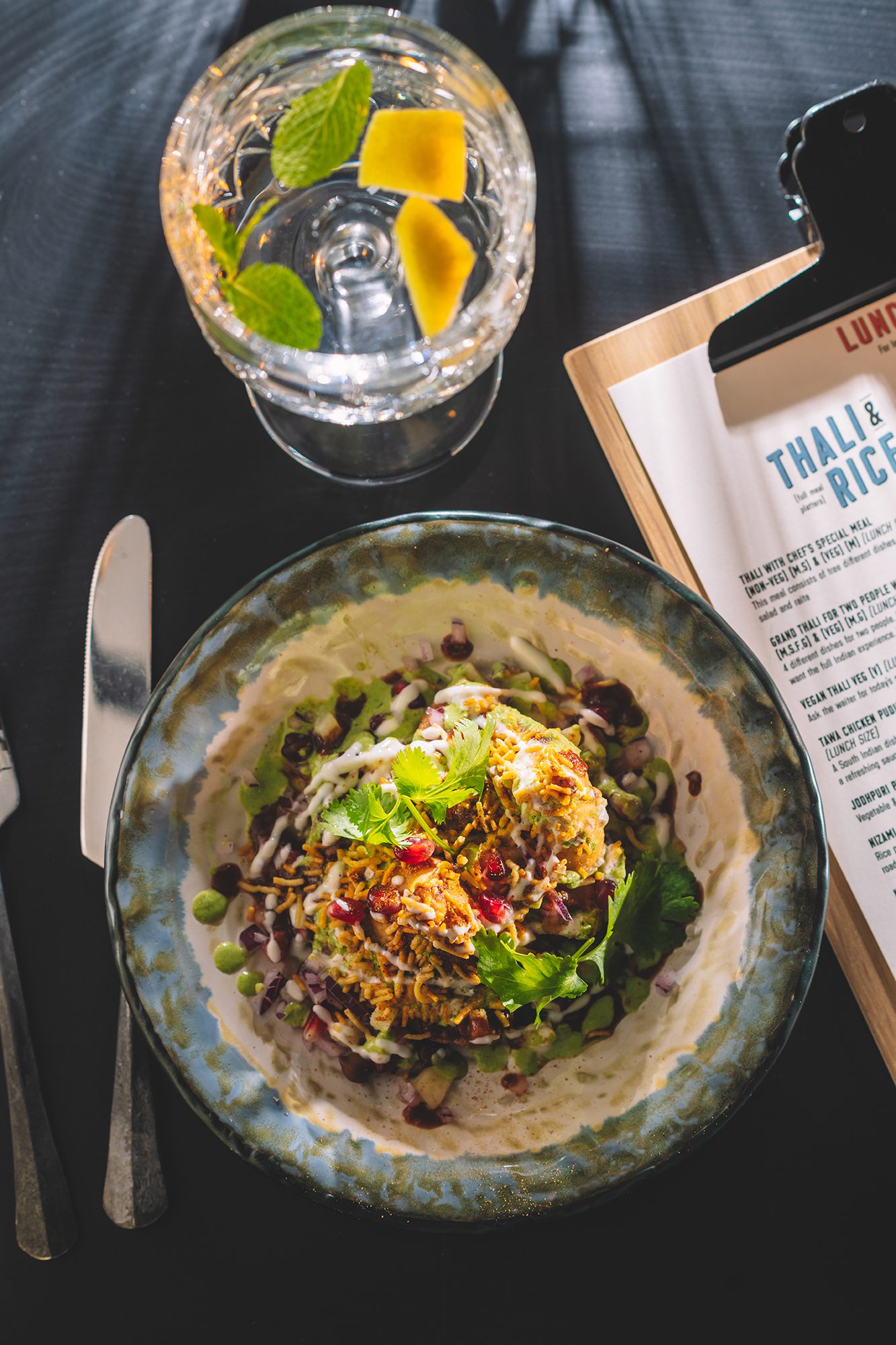 Samosa chaat på bildet med ulike farger, sauser, graneteple og korriander. Vann med sitron og korriander på bordet. Menyen på bordet. Kniv på bordet. Solstråler i bildet.