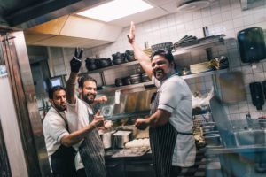 3 kokker som smiler mens de lager mat. En viser tommelopp, en viser "peace" tegn. I bakgrunnen har du naan-deig og ulike skåler til maten.
