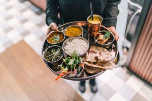 En servitør som holder thali, som er en tallerken med mange ulike matretter og mango lassi. Servitøren har på svart unfirom.