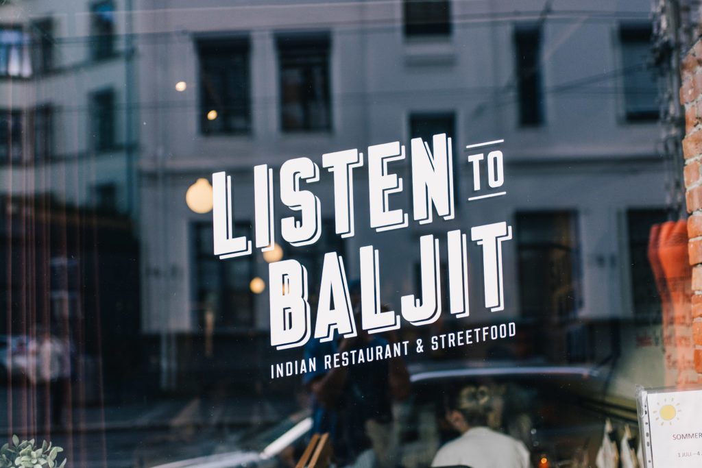Hvit "Listen to Baljit" logo på vinduet til restauranten. I speilbildet ser man et hvitt bygg.