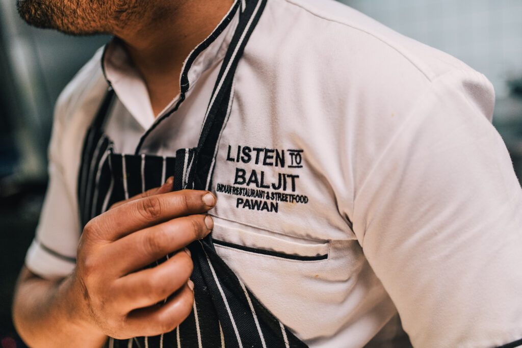 Kokk med forkle viser logo for Listen to Baljit på skjorten